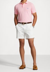 Rosa polo skjorta med en liten blå logotyp, vita shorts, brun bälte och bruna loafers. Modellen poserar med händerna i fickorna.