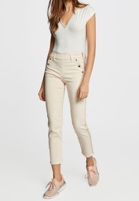Jeans de tiro alto en color beige claro con botones frontales y cremallera lateral, combinados con una camiseta ajustada de cuello en V blanca y zapatillas en tono rosa empolvado.