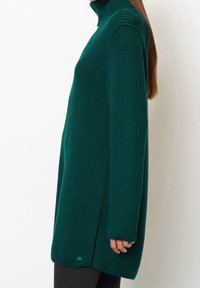 Rollkragenpullover in tiefem Teal, mit gerippter Textur, seitlichen Schlitzen und langen Ärmeln. Ein kleines Markenetikett ist am Saum sichtbar.