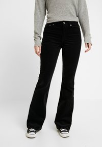 Svarta utsvängda kordbyxor med hög midja, som har fem fickor, knappstängning och en subtil ribbad textur, tillsammans med svarta sneakers.