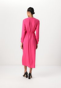 Fuchsia long-sleeve dress with a cinched waist, flowing design, and smooth texture. Paired with black block-heeled shoes.