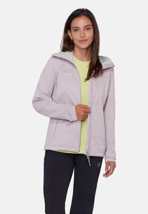 Vrouw draagt een lichtpaarse jas met rits over een geel shirt en zwarte broek, staand tegen een effen witte achtergrond.