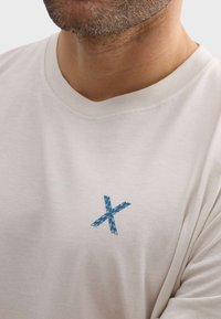 Camiseta de algodón color beige claro con bordado de un patrón de hojas azules en forma de X en la parte izquierda del pecho, cuello redondo y textura suave.