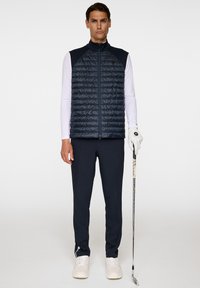 Marinblått quiltat väst med hög krage, bärs över en långärmad vit skjorta, i kombination med mörka byxor och vita sneakers, hållande en golfklubba.