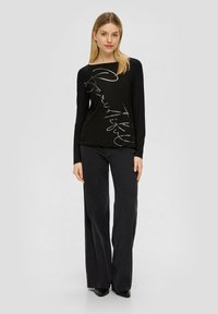 Czarny top z długim rękawem z metalicznym srebrnym, ręcznie napisanym tekstem. Połączony z szerokimi czarnymi spodniami i czarnym obuwiem. Gładka tekstura tkaniny.