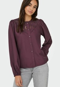 Jonge vrouw in een pruimenblauwe blouse met knopen en kantdetails op de kraag, in combinatie met lichtgrijze jeans, staand met één hand in de zak.