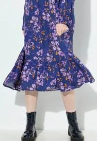 Lila blumiges Midikleid mit orangefarbenen Akzenten, das einen gerafften, gestuften Saum aufweist. Kombiniert mit schwarzen, patentierten Schnürstiefeln.
