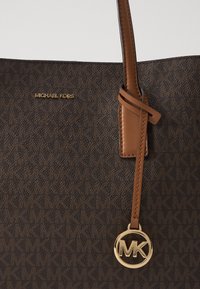 Barna designer vászontáska monogram mintával, bőr részletekkel, arany tónusú hardware-rel és lógnak a táska oldalán egy MK logó charm.