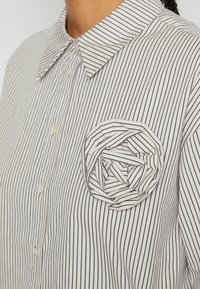 Close-up of white button-up shirt with thin vertical black stripes and a fabric rose detail on the chest pocket area.