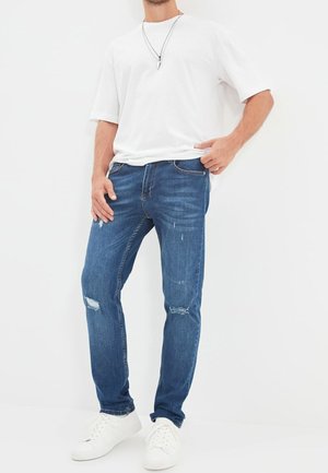 Man draagt een wit T-shirt, blauwe gescheurde spijkerbroek, witte sneakers en een ketting, staand tegen een witte achtergrond.