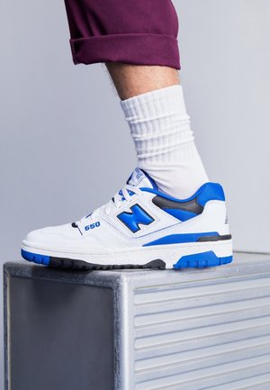 Pied portant une basket New Balance 550 blanche et bleue avec une chaussette blanche et un pantalon violet retroussé, reposant sur une plateforme métallique.