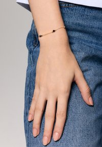 Złota bransoletka z łańcuchem, z dwoma okrągłymi czarnymi koralikami, noszona na ręce opierającej się na dżinsie. Gładka faktura, delikatny design i minimalistyczne wykończenie.