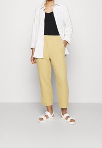 Pantalon texturé jaune clair avec une taille élastique, associé à un débardeur noir et une chemise blanche à boutons. Des sandales blanches complètent le look.