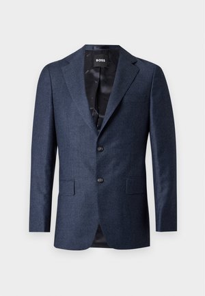 Homme portant un costume bleu foncé, chemise blanche, chaussures noires, tenant une mallette noire, debout devant un fond clair uni.