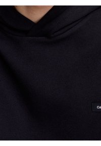 Sudadera negra de tela suave, con capucha ajustable y una pequeña etiqueta de marca en la parte delantera. Textura suave, diseño minimalista.