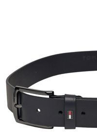 Cintura in pelle nera con una fibbia in metallo nero lucido. Un piccolo logo rosso, bianco e blu è posizionato vicino alla fibbia.