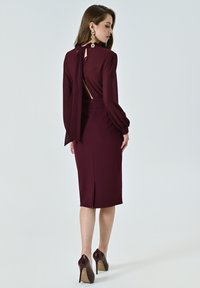 Abito fitted bordeaux con maniche lunghe, design con schiena scoperta e dettaglio al collo legato. Tessuto liscio con spacco posteriore e abbinato a tacchi alti.
