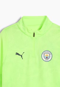 Vihreä, puoliveto-ketjuvarustus, joka on valmistettu tekstuuripinnoitteisesta kankaasta, jossa on musta Puma-logo ja Manchester City -merkki rinnassa.