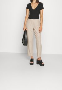 Svart kortärmad bodysuit med spetsdetaljer, beige skräddarsydda byxor och svarta klacksandaler. Modell som håller en svart väska.