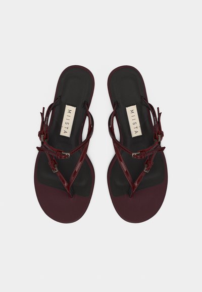 Ζευγάρι σκούρα κόκκινα φλατ σανδάλια με λεπτά λουράκια και αγκράφες, τετράγωνη μύτη και μαύρη σόλα με ετικέτα «MIISTA», απεικονιζόμενα από πάνω.