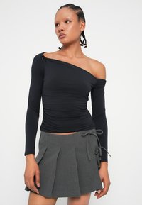 Crna bluza dugih rukava s jednim ramenom otkrivenim, s naboranim dizajnom. Uparena s sivom plisiranom suknjom i mašnom na struku.