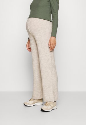 Personne portant un pantalon beige large côtelé, des baskets beiges et un haut à manches longues vert olive, debout devant un fond clair uni.