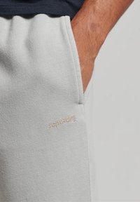 Ruka osoby v kapse světlých šedých tepláků s vyšitým logem "superdry" blízko okraje kapsy.