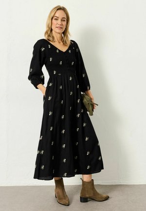 Woman in a black midi dress with small floral prints, puffed sleeves, holding an olive green clutch, wearing brown suede ankle boots.