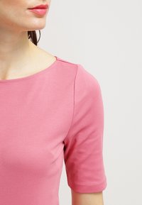 Růžová košile s krátkým rukávem a kulatým výstřihem, vyrobená z hladkého, elastického materiálu. Obsahuje jemné švy na ramenou.