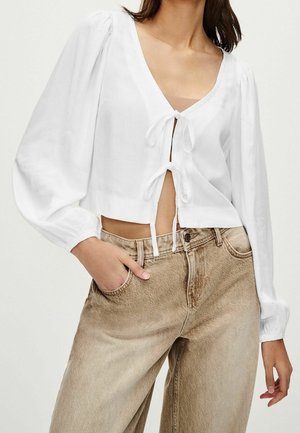 Vrouw draagt een wit cropped blouse met lange pofmouwen en een strik aan de voorkant, gecombineerd met een beige high-waisted jeans, hand in de zak.