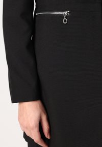 Svart plagg med en slät textur, som har en horisontell ficka med dragkedja vid midjan och en silverfärgad dragkedjehandtag som accent.