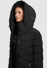 Jachetă puffer neagră cu guler înalt și glugă supradimensionată, având o textură matlasată, închidere cu fermoar și un accent de logo pe manșetă.