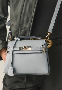 Világoskék, texturált bőrtáska arany csattal és levehető pánttal, keresztben viselve fekete ruházat és olívazöld dzseki felett.