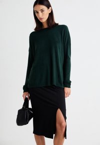 Pulover lung de culoare verde închis, cu mâneci lungi, asortat cu o fustă tip creion, texturată, de culoare neagră, cu o fanta laterală. Modelul ține o poșetă mică de culoare neagră.