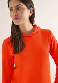 Femme aux longs cheveux bruns portant un sweat à capuche côtelé orange vif, regardant sur le côté avec un léger sourire sur un fond clair.