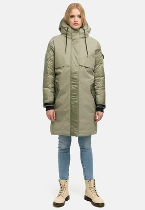 Jeune femme portant un long manteau matelassé vert olive avec capuche, un jean skinny bleu et des bottines beige lacées, debout devant un fond uni.