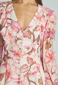 Vestido floral con escote en V, que presenta patrones de flores rosas y verdes, mangas con volantes y detalles de botones en una tela suave y ligera.