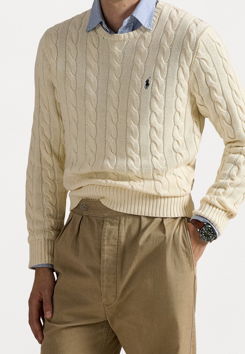 Kremowy sweter w warkocze z prążkowanym obszyciem i mankietami, noszony na jasnoniebieskiej koszuli; zestawiony z beżowymi spodniami i ciemnym zegarkiem na nadgarstku.