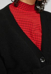 Cárdigan negro con diseño de pico, que presenta un botón brillante y una textura suave, superpuesto sobre un cuello alto rojo con un patrón a rayas negras.