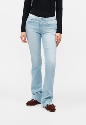 Personne portant un jean bootcut bleu clair, un haut noir moulant à manches longues et des chaussures marron à lacets blancs, debout devant un fond uni.
