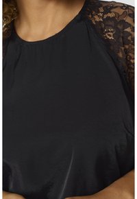 Blouse noire avec manches en dentelle florale noire transparente et col rond sur une personne aux cheveux bouclés.
