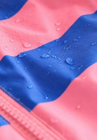 Waterafstotende stof met gedurfde roze en blauwe strepen. Gelaagde oppervlakte met waterdruppels; zichtbare rits langs de rand.