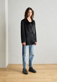 Svart dobbeltradet blazer med glatt tekstur, kombinert med lyseblå denimjeans og svarte ankelstøvler, satt mot en nøytral bakgrunn.