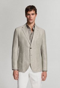 Homme portant un blazer en lin gris clair sur une chemise grise et un pantalon blanc, debout devant un fond clair uni.