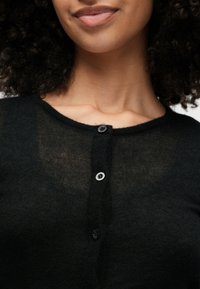 Črn, lahek cardigan z okroglim izrezom, tremi gumbi in prozorno teksturo. Tkanina se zdi mehka in rahlo prilegajoča ob izrezu.