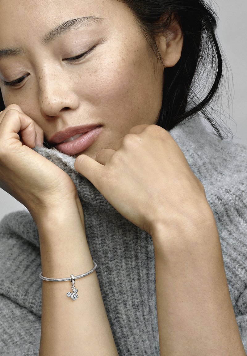 Srebrna zapestnica s charmom ima tri prozorne akcentne gemstone. Nosilka je oblečena v teksturirano sivo pleteno jopico, z rokami blizu obraza.