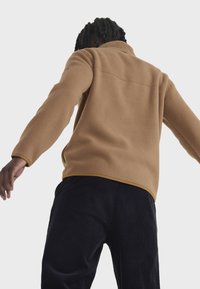 Beige Fleecejacke mit hohem Kragen und gelben Akzenten, langen Ärmeln und weichem Material, kombiniert mit dunklen Cordhosen.