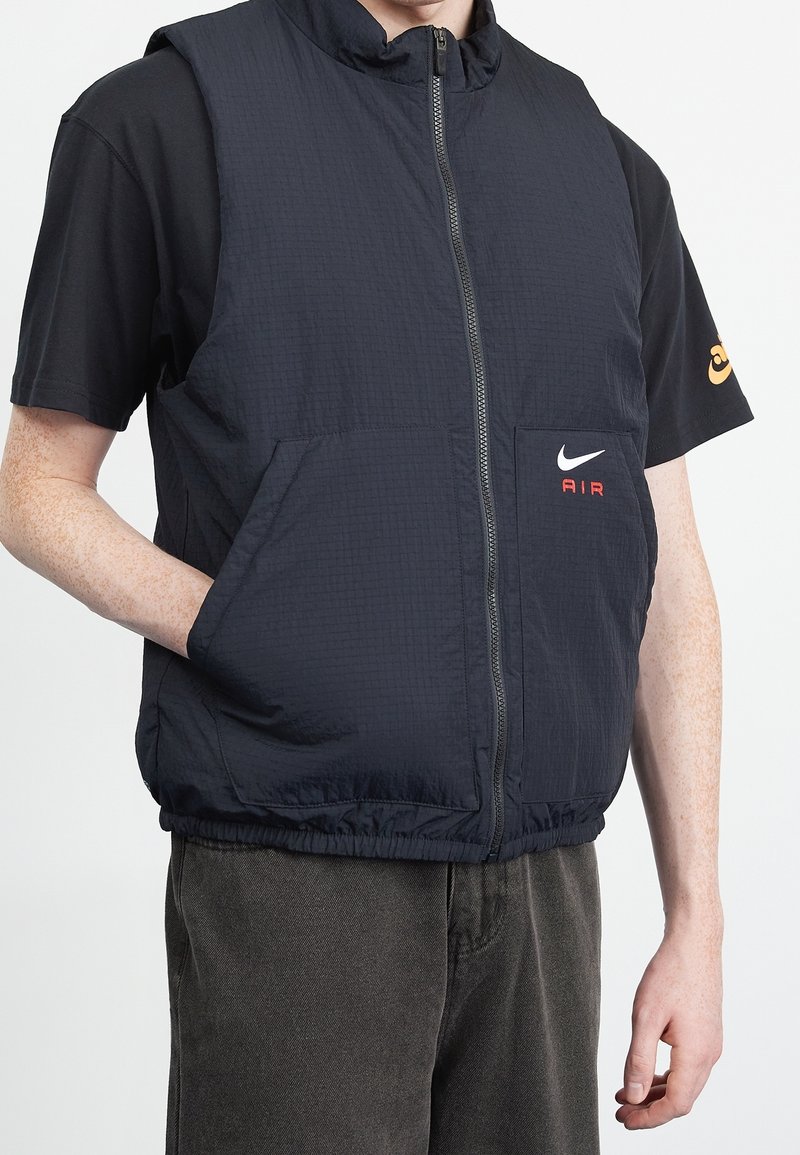 Svart lättviktsväst med hög krage, framdragkedja och Nike-logotyp. Har två framfickor och elastisk fåll. Bärs över en svart t-shirt.