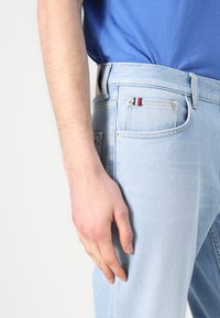 Mies, jolla on vaaleansiniset farkut ja sininen paita, käsi lepää reiden päällä pienen taskun lähellä, jossa on punainen, valkoinen ja sininen lappu.