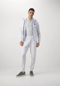 Man staat met een witte ritssluiting hoodie, witte jogger broek, lichtgrijs polo shirt en grijze sneakers tegen een effen achtergrond.